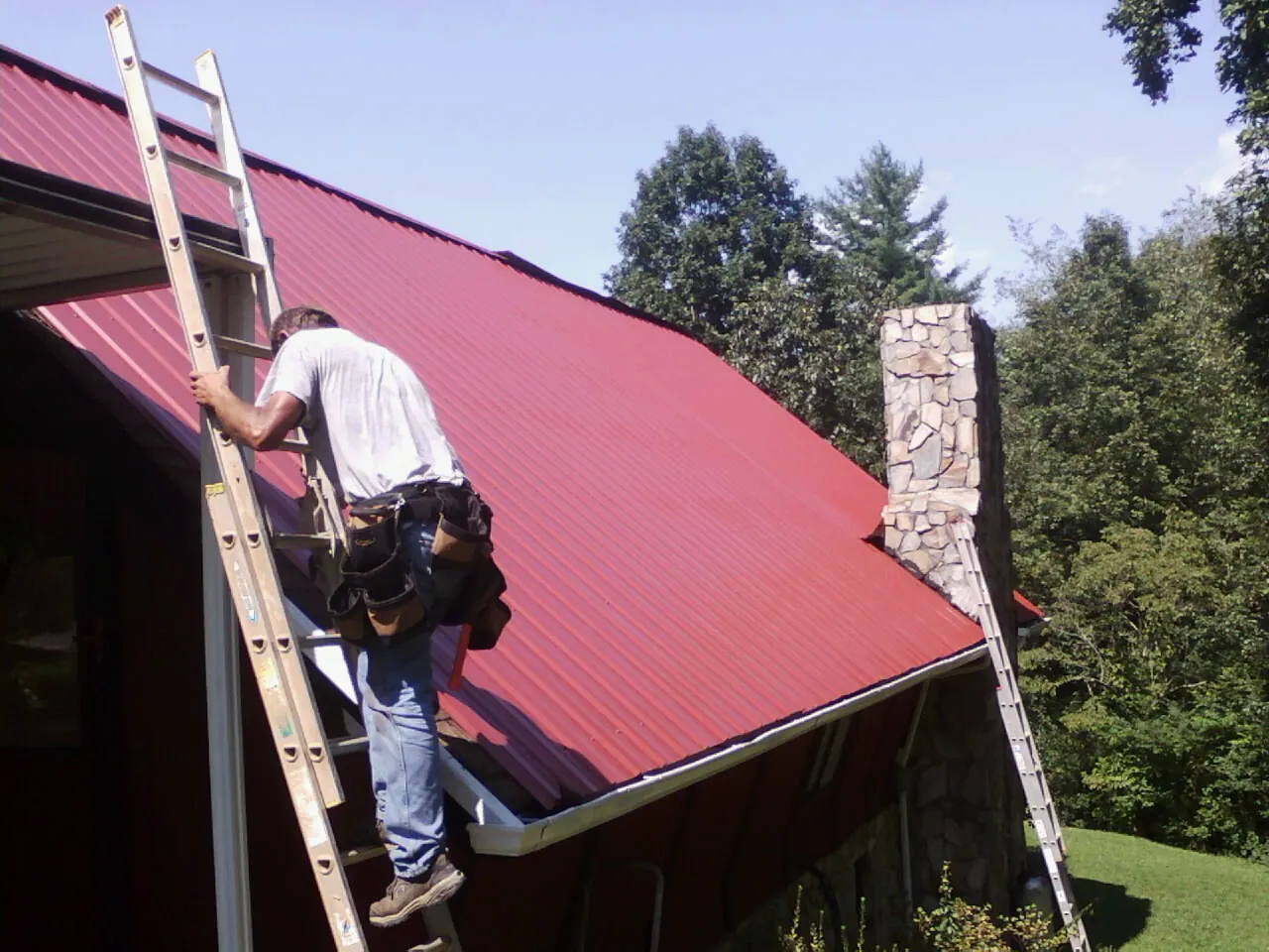 Roofer on a ladder beside a red metal roof and stone chimney in Hillpoint